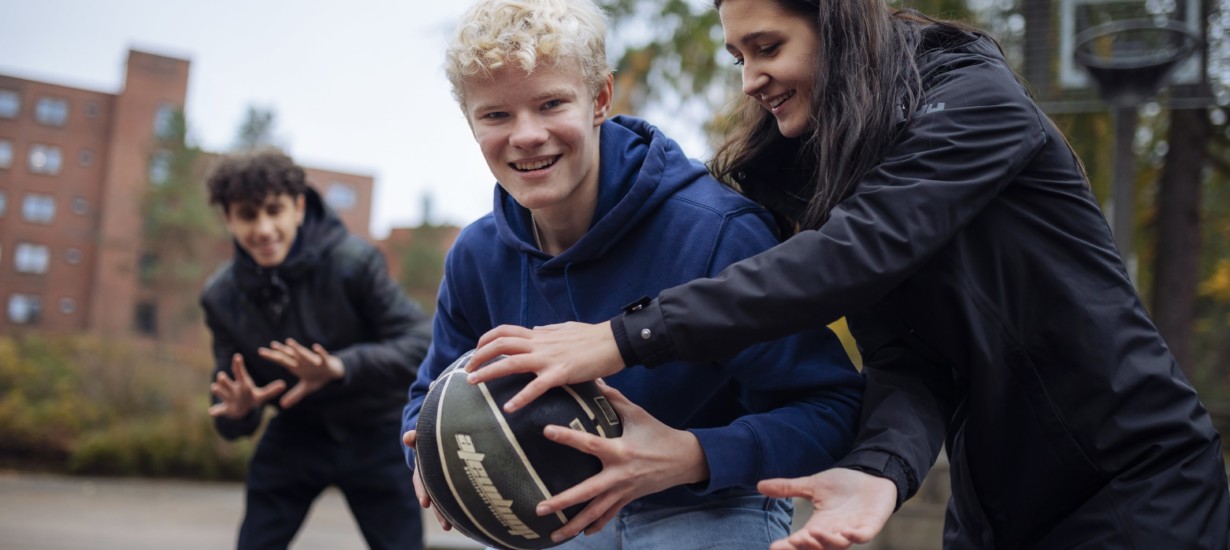 Kolme nuorta pelaa koripalloa ulkona.