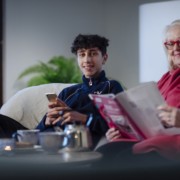 Isoäiti ja nuori poika istuvat sohvalla. Isoäiti lukee lehteä ja poika katsoo kameraan ja hymyilee.