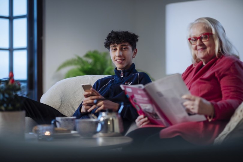 Isoäiti ja nuori poika istuvat sohvalla. Isoäiti lukee lehteä ja poika katsoo kameraan ja hymyilee.