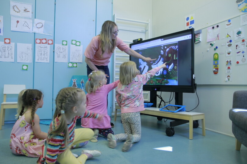 Lapset ja varhaiskasvatuksen opettaja osoittavat televisioruudulla olevaa virtuaalimaailma Rauhametsää.
