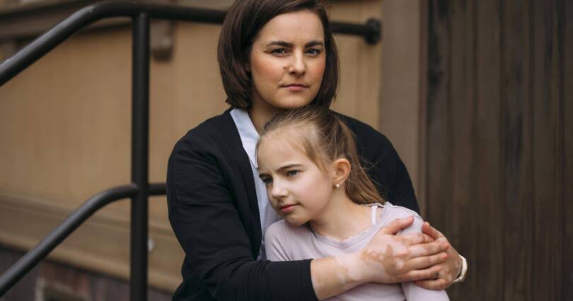 Vanhempi pitää lasta sylissään. Taustalla portaan kaide ja talon seinä, jossa ikkuna.