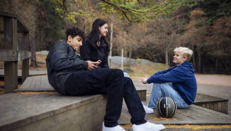 Kolme nuorta ulkona istumassa koripallo mukana