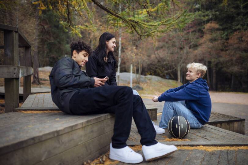 Kolme nuorta ulkona istumassa koripallo mukana