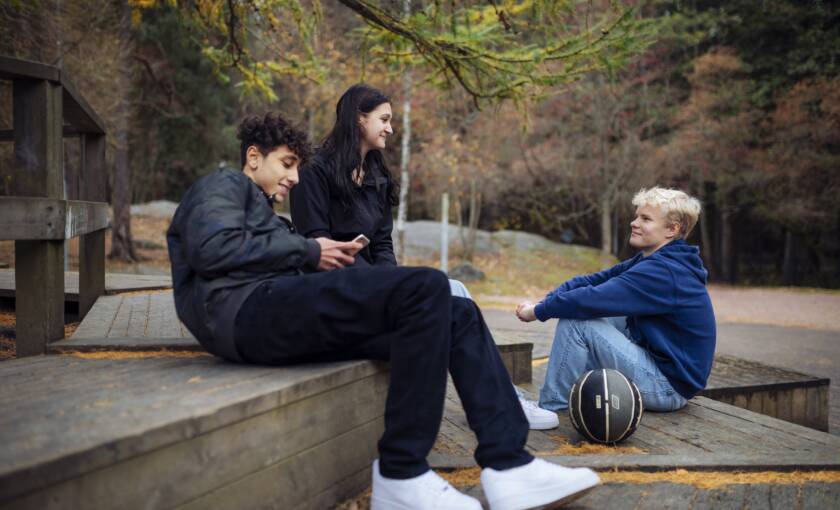 Kolme nuorta ulkona istumassa koripallo mukana