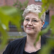 Kirjailija Raisa Omaheimo katsoo kuvassa suoraan kameraan. Vihreät lehdet reunustavat kuvaa. Kuva on otettu Länsi-Pasilassa Helsingissä.