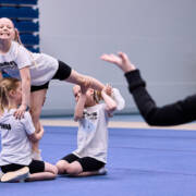 Kolme pientä Cheerleadingin harrastajaa tekevät kuviota ja etualalla mustiin puekutunut valmentaja ohjaa käsillään.