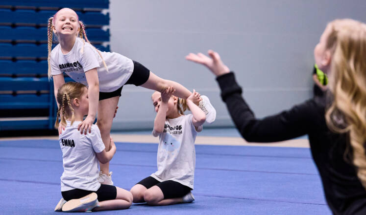Kolme pientä Cheerleadingin harrastajaa tekevät kuviota ja etualalla mustiin puekutunut valmentaja ohjaa käsillään.
