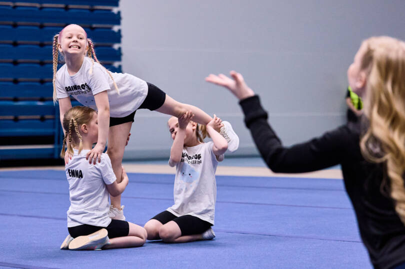 Kolme pientä Cheerleadingin harrastajaa tekevät kuviota ja etualalla mustiin puekutunut valmentaja ohjaa käsillään.