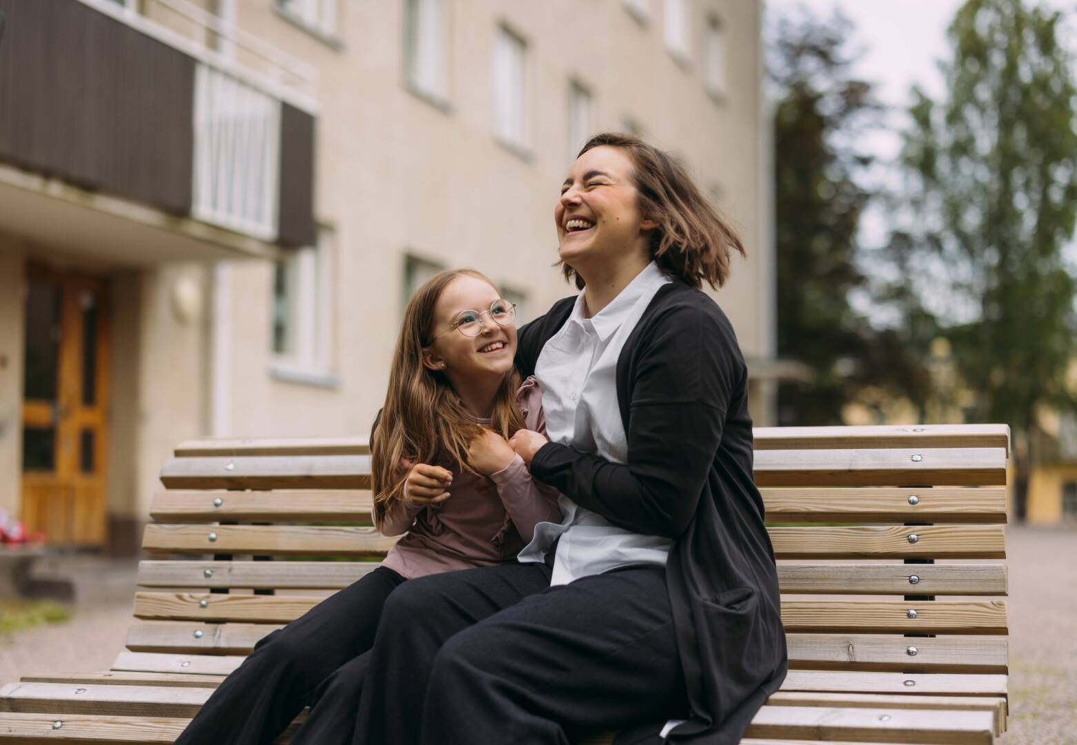 Nainen ja lapsi istuvat puiston penkillä. Aikuinen halaa lasta ja nauraa.