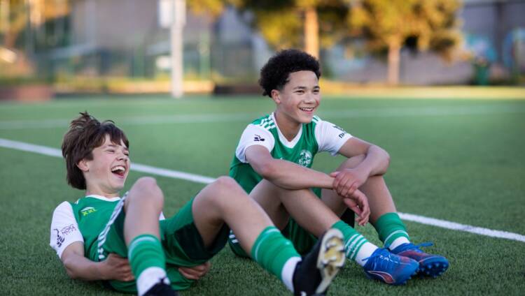 Kaksi nuorta jalkapallon pelaajaa istuu kentällä ja hymyilee