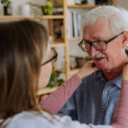 Naisoletettu takaapäin pitää käsiä valkohapsisen miehen hartioilla. Olisiko vaikapa hoitajan ja palvelutalon asiakkaan kohtaaminen?