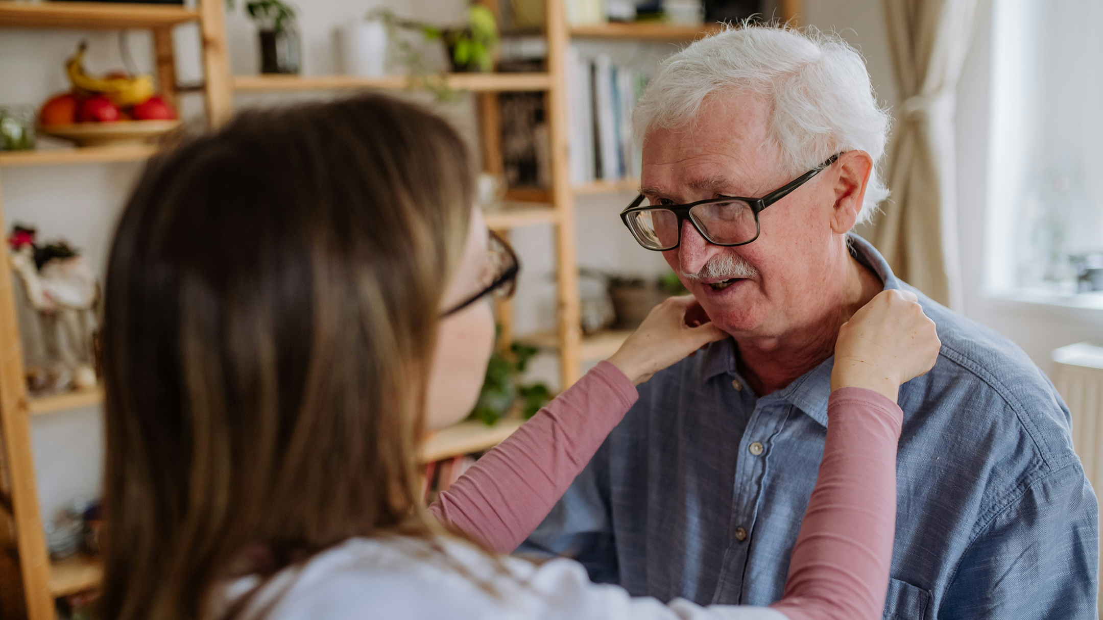 Naisoletettu takaapäin pitää käsiä valkohapsisen miehen hartioilla. Olisiko vaikapa hoitajan ja palvelutalon asiakkaan kohtaaminen?