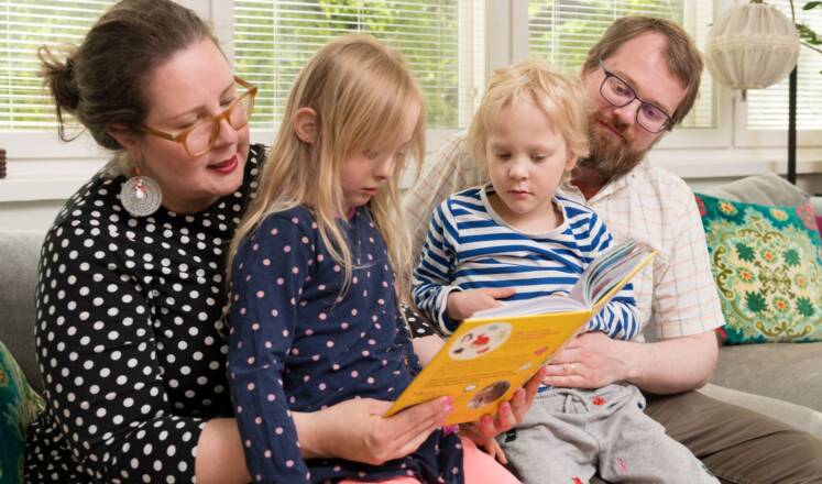 Perhe istuu sohvalla. Kaksi alakouluikäistä lasta lukee kirjaa vanhempien sylissä.