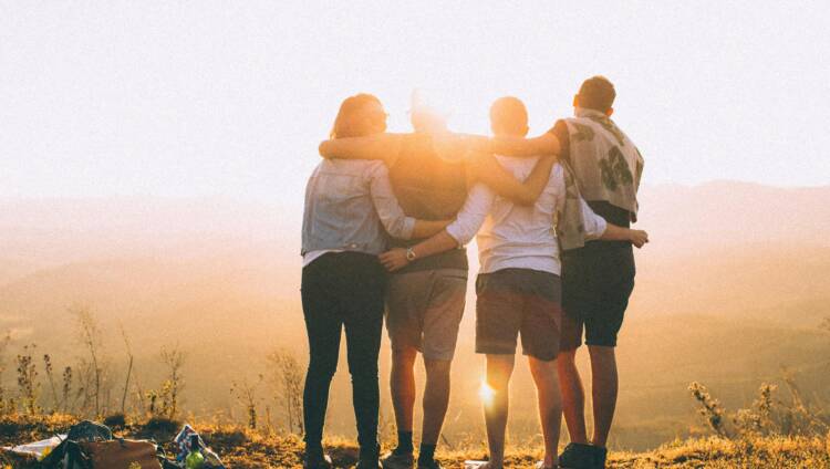Neljä ihmistä kädet toistensa harteilla ihailemassa tunturimaisemassa auringonnousua, takaapäin kuvattuna.
