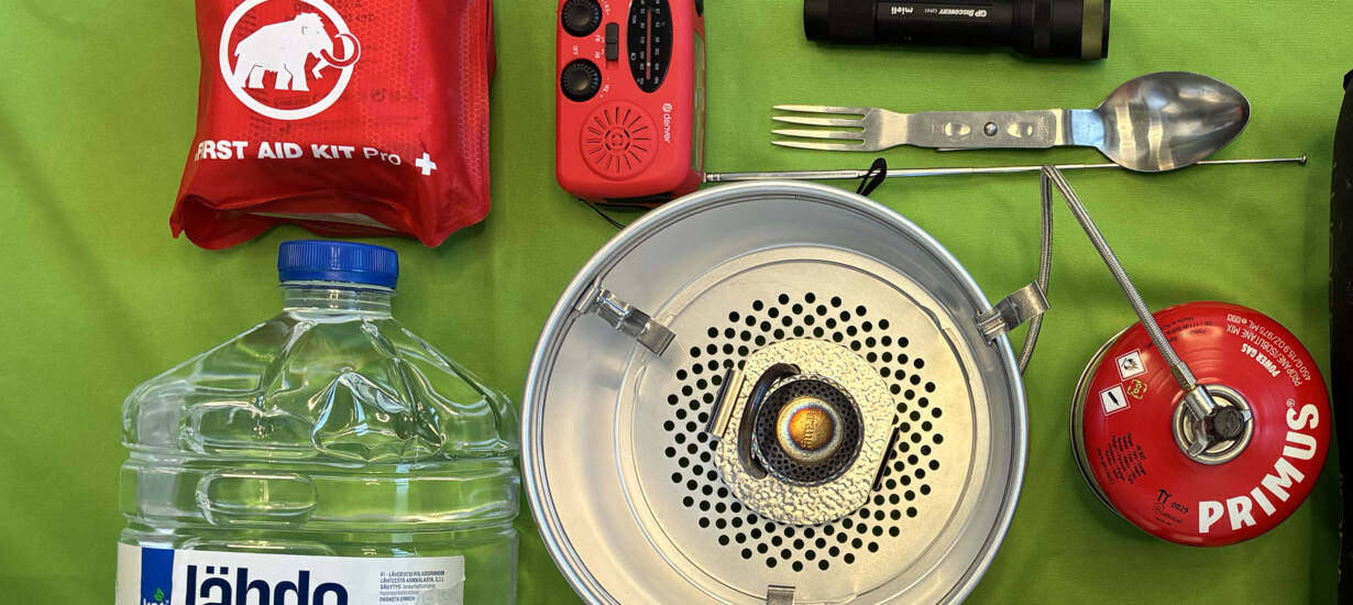 Vihreällä taustalla välttämättömyystarvikkeita: ensiapupakkaus, patteriradio, trangia, spork, makuupussi.